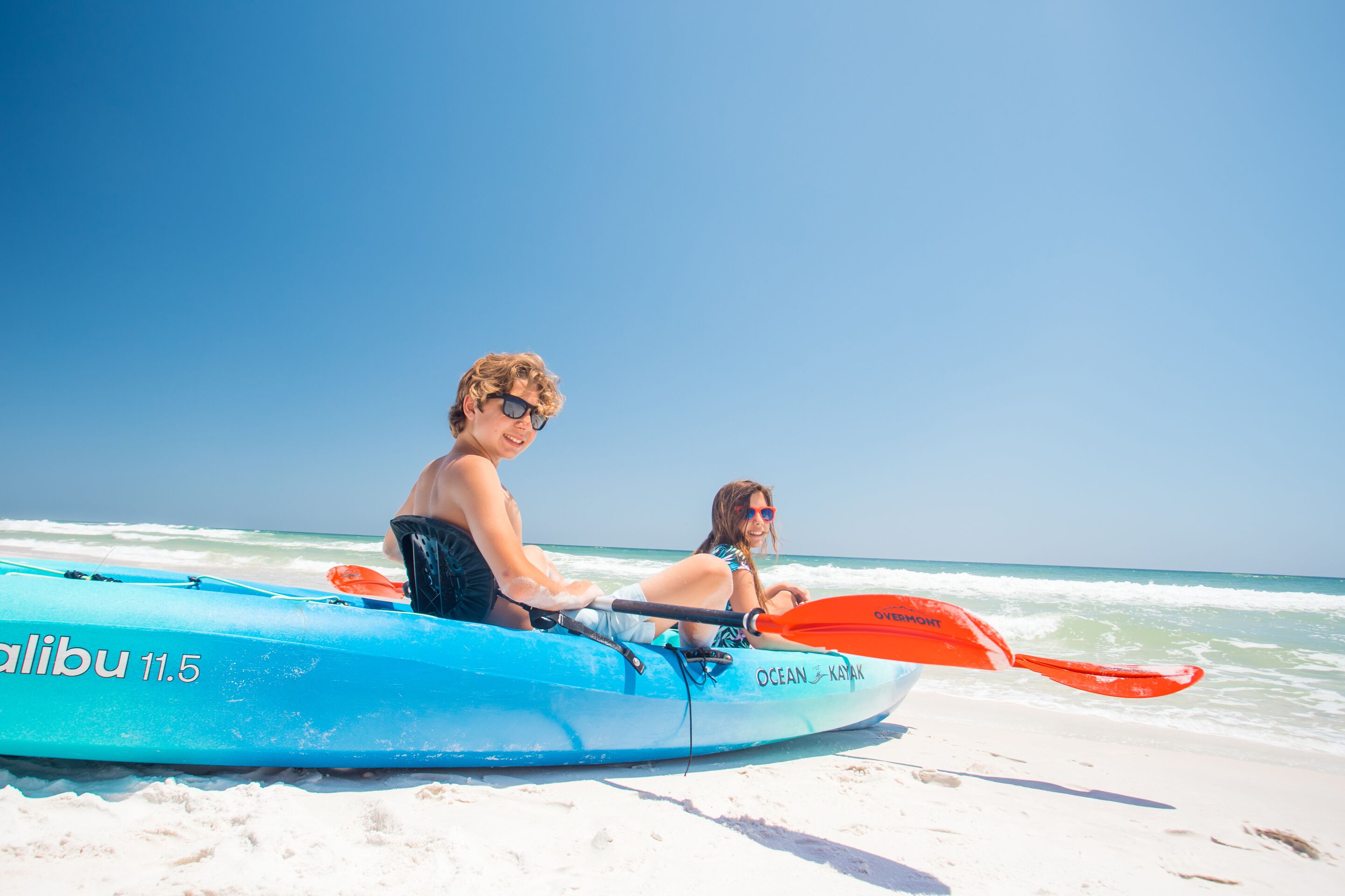 Ein blaues Kajak mit der Aufschrift "Ocean Kayak" und "Alibu 11.5", das auf einem sandigen Strand liegt, mit dem Meer im Hintergrund.