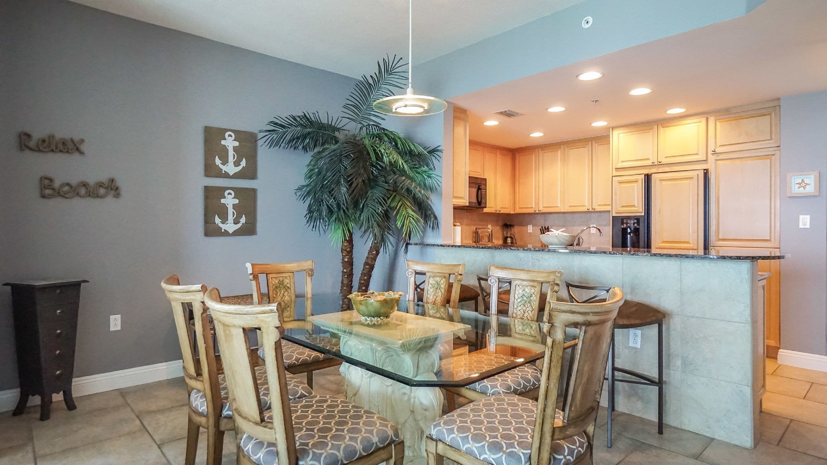 A dining area with a glass table and wooden chairs, a kitchen with wooden cabinets, and a decorative plant.