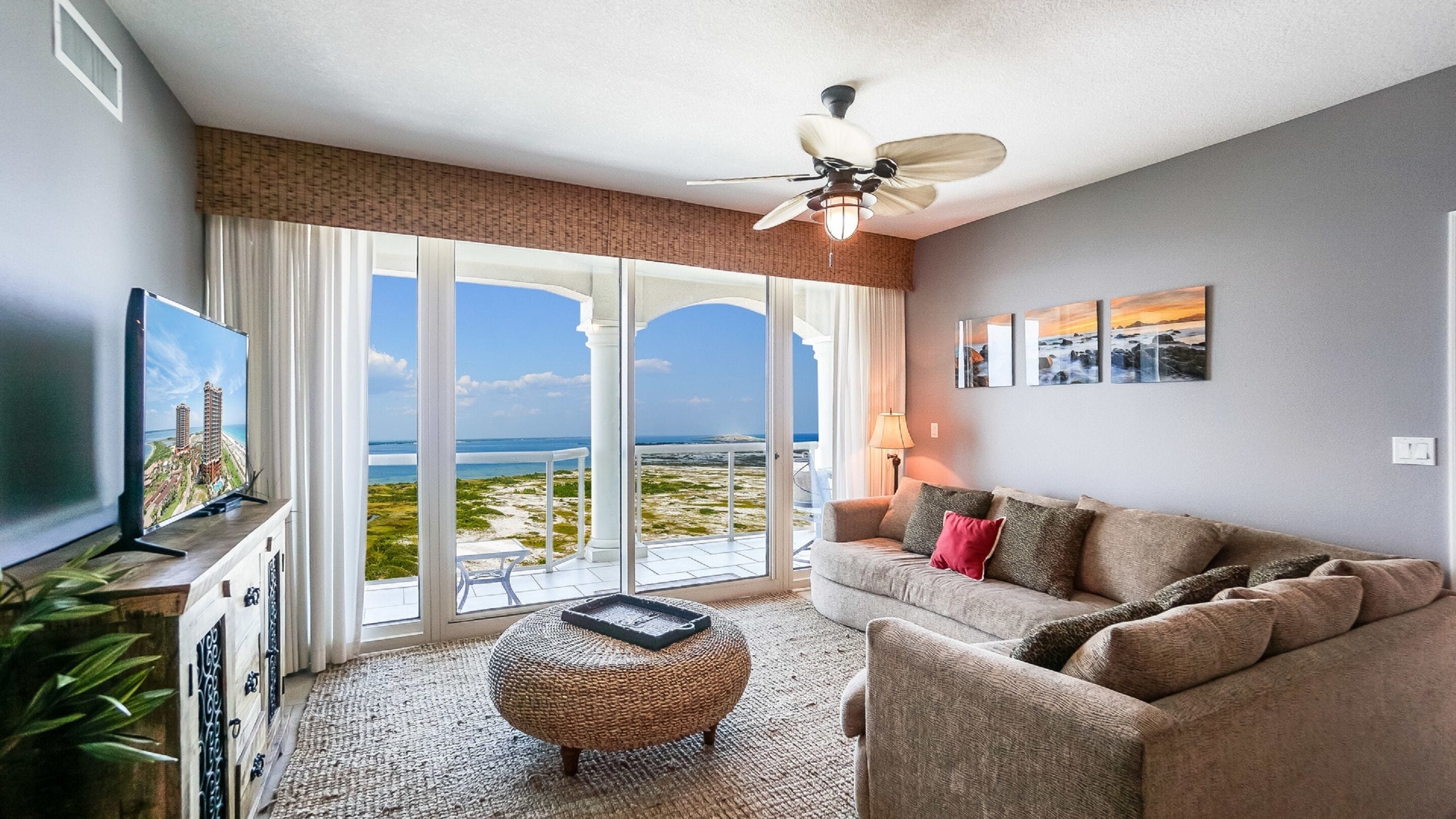 A living room with a large TV, a sofa, a coffee table, and a view of the beach through large glass doors.
