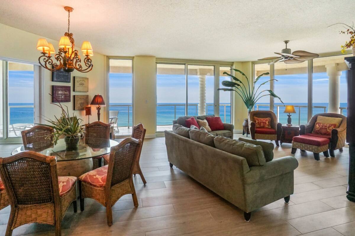 A spacious living room with a large window overlooking the ocean, a chandelier, and a dining area with wicker chairs.