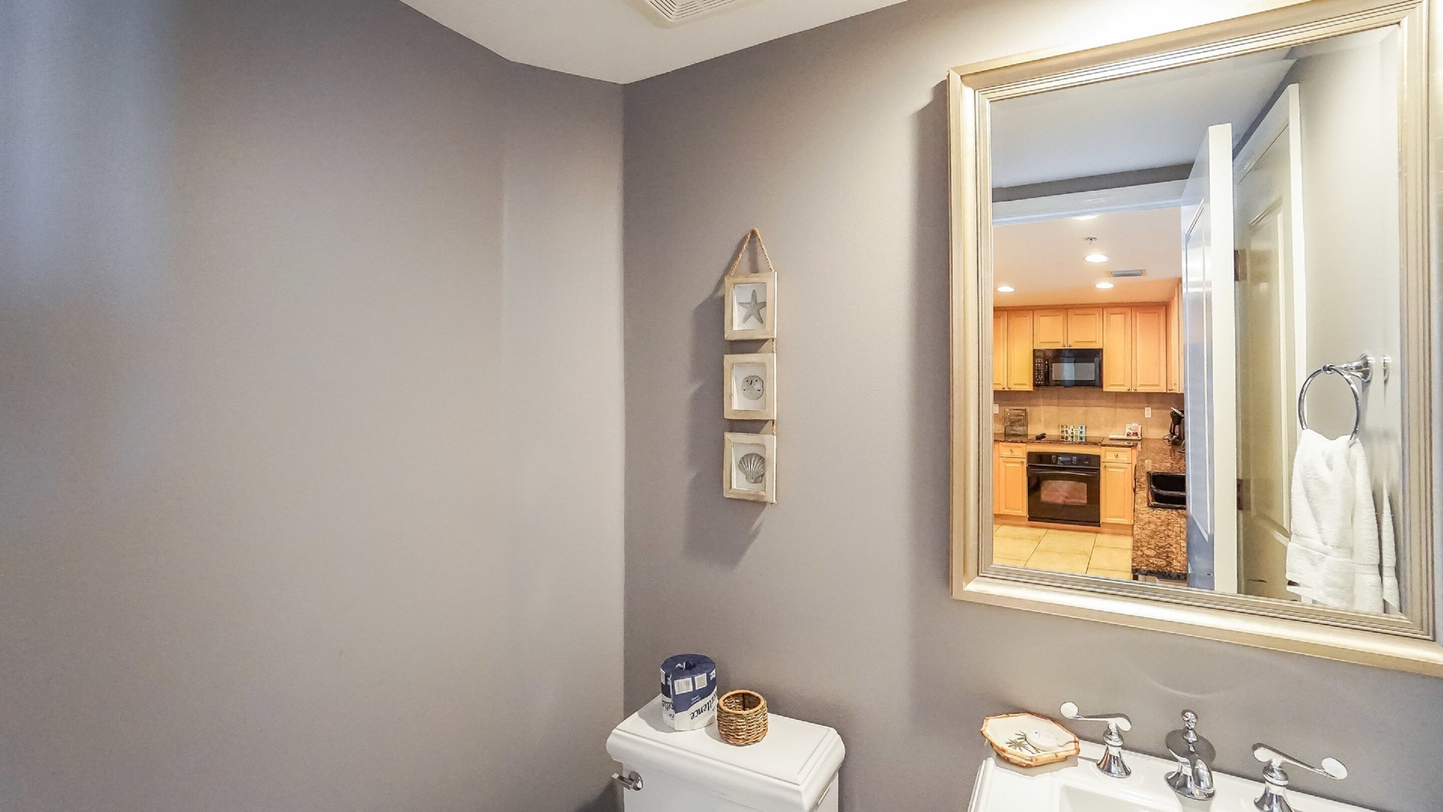 A bathroom with a toilet, sink, and a large mirror reflecting a kitchen area.