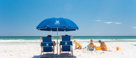 Een strand met een blauwe parasol en twee blauwe stoelen met een logo, zandstrand en de oceaan op de achtergrond.