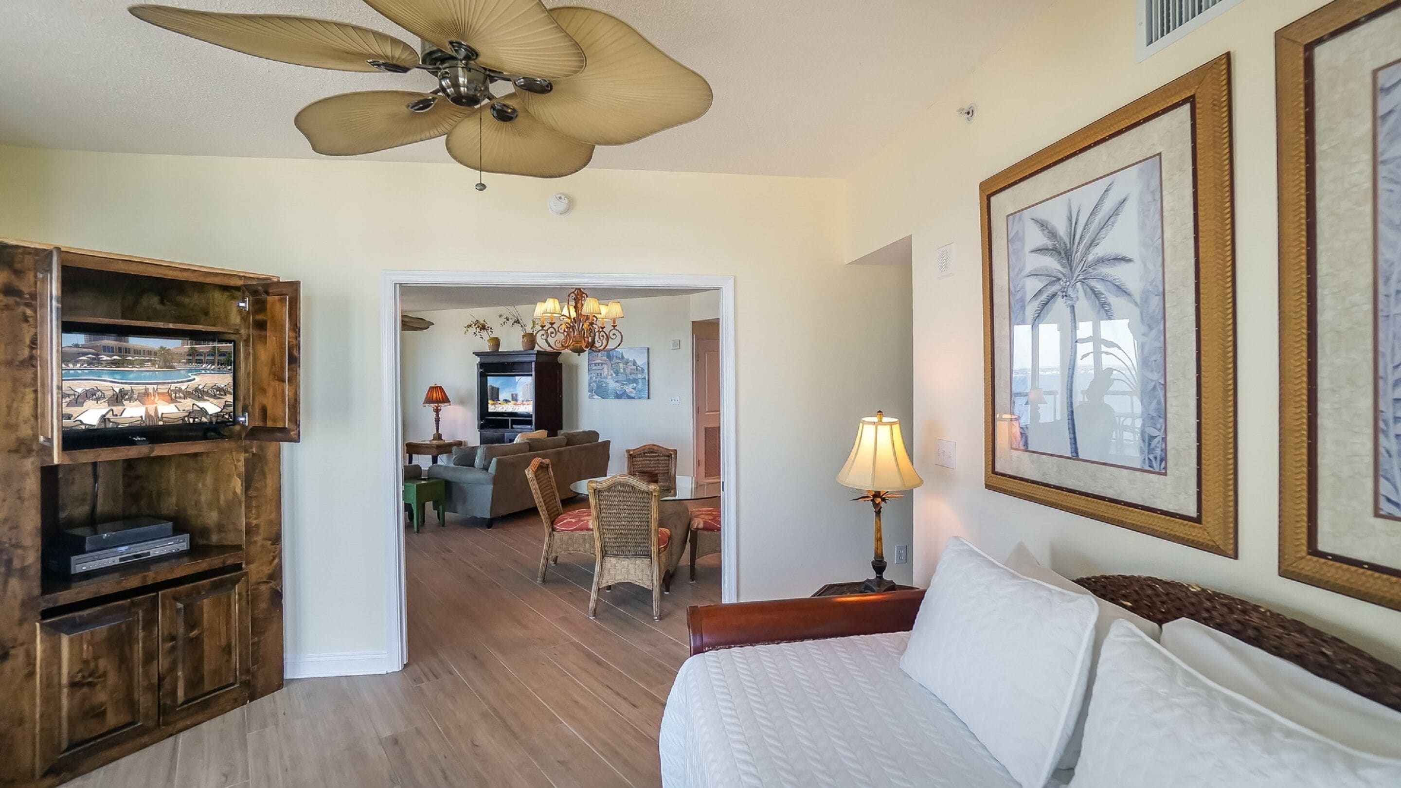 A hotel room with a bed, a ceiling fan, a TV, and a dining area with a chandelier.