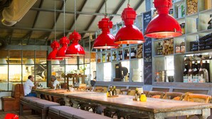 Un restaurant avec une longue table en bois, des lampes suspendues rouges et un coin bar avec des étagères remplies de bouteilles.