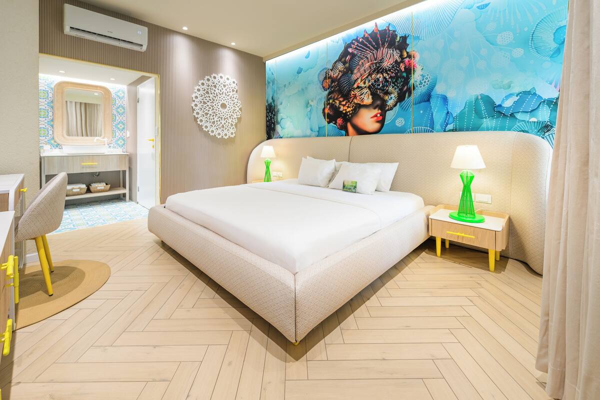 Une chambre d’hôtel moderne dotée d’un grand lit, de deux tables de chevet avec lampes, d’une fresque murale décorative et d’un parquet en bois à motifs chevrons.