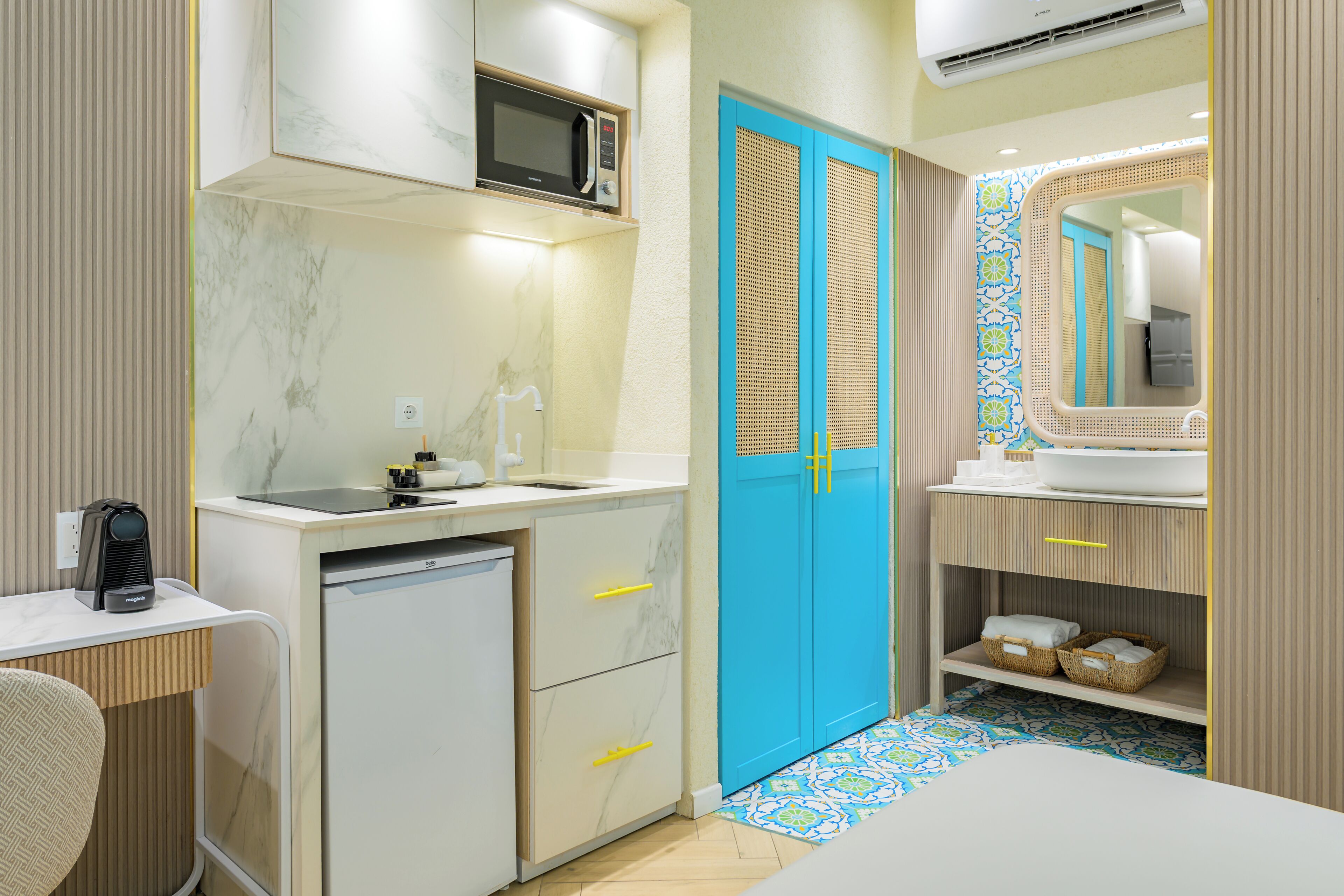 Habitación de hotel compacta con kitchenette, microondas, lavabo, refrigerador pequeño, puerta azul, espejo y piso con diseño.