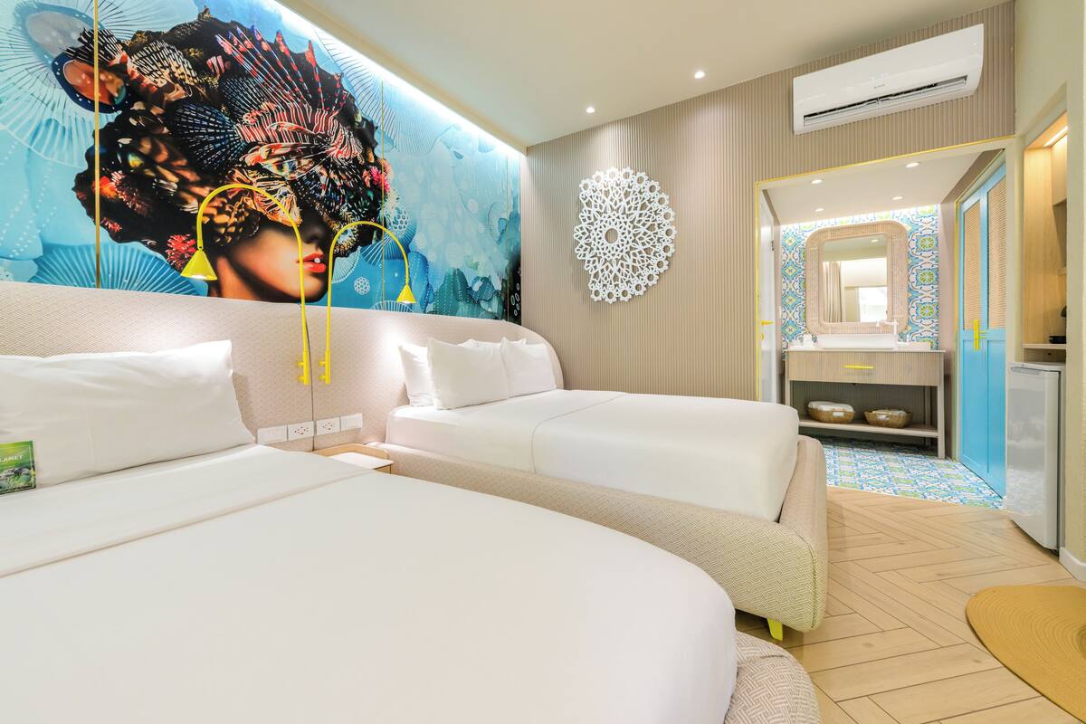 Une chambre d’hôtel avec un grand lit, un canapé, une fresque murale décorative, une salle de bain avec un miroir et un lavabo, et un sol à motifs.