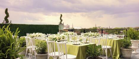 An outdoor dining area with tables set for a meal, surrounded by potted plants and white chairs.