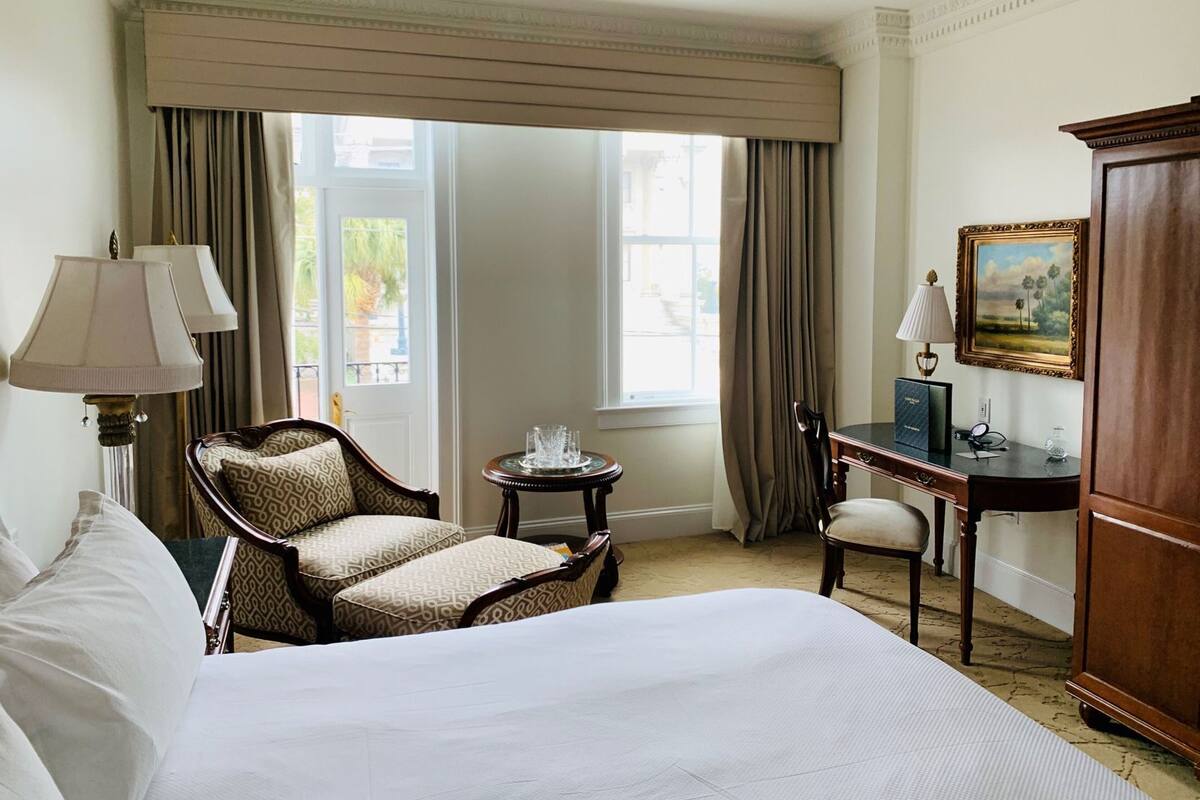 A hotel room with a large bed, a desk, a chair, a lamp, a cabinet, and a painting on the wall.