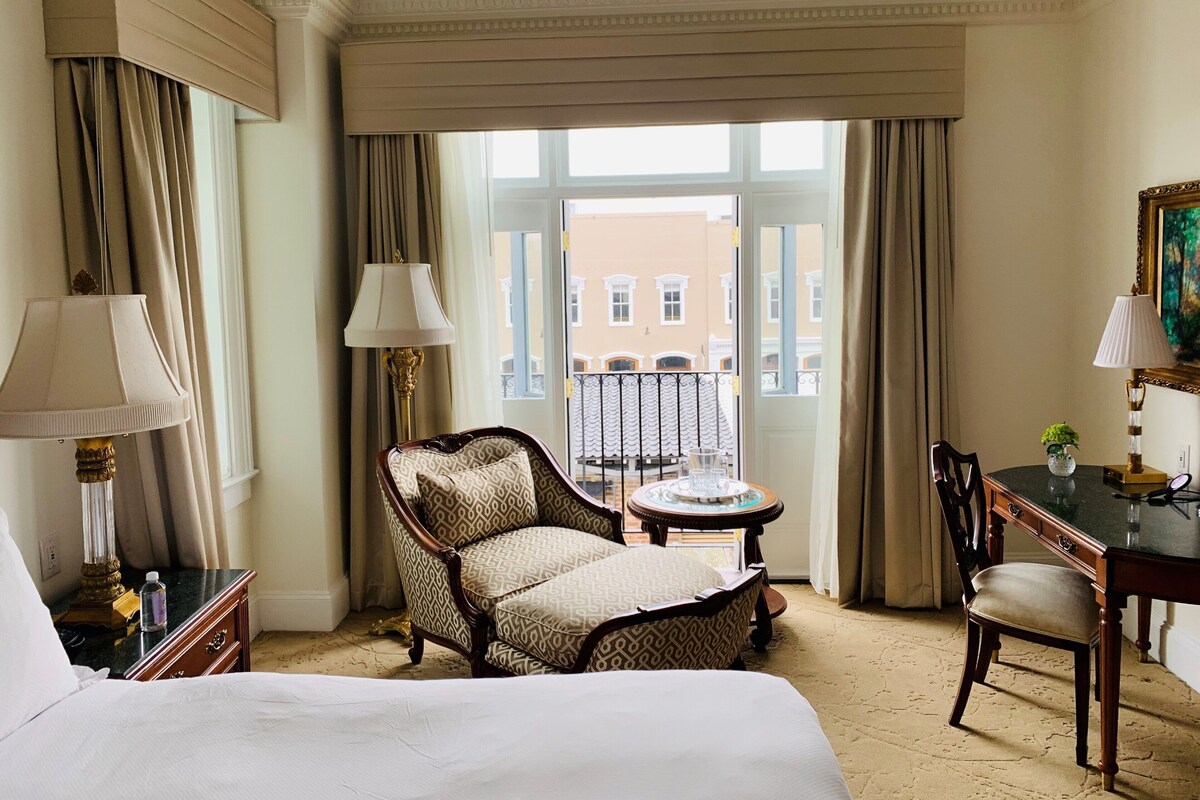 Quarto de hotel com cama, duas cadeiras, uma mesinha e uma escrivaninha com abajur.