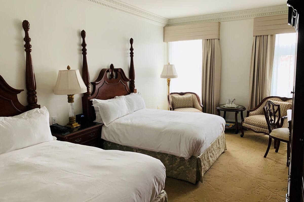 A hotel room with two beds, a chair, a small table, and a lamp.