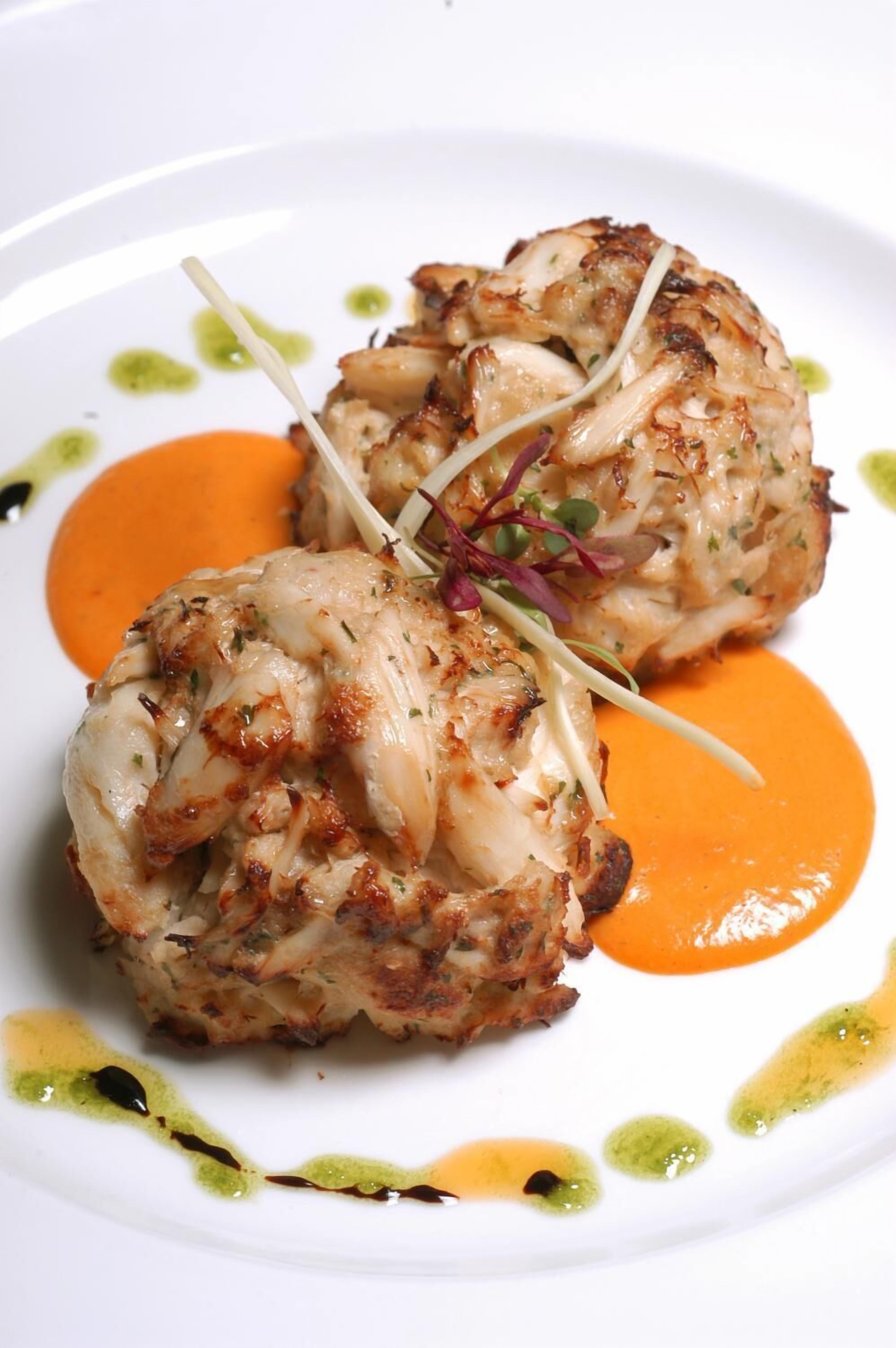 A plate with two crab cakes, garnished with microgreens and toothpicks, served with a side of orange sauce.