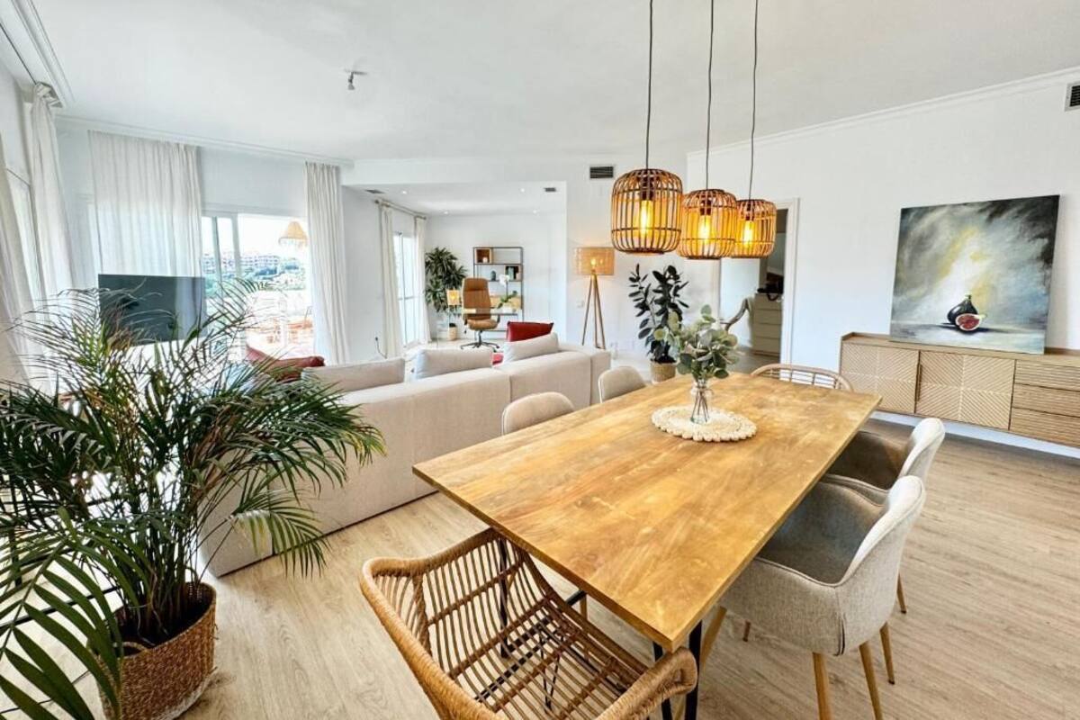 A spacious living room with a wooden dining table, wicker chairs, a large potted plant, and a modern chandelier.