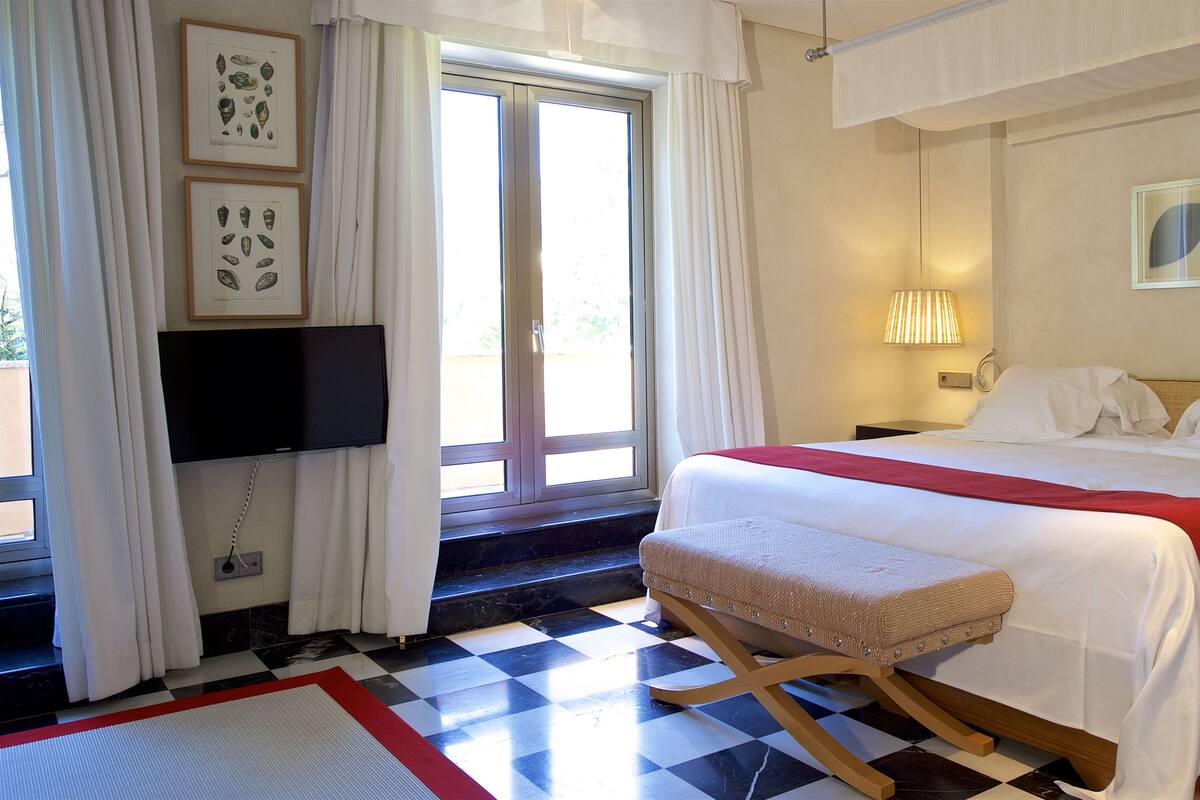 A hotel room with a checkered floor, a bed with white bedding and a red accent, a television on the wall, and a window with curtains.