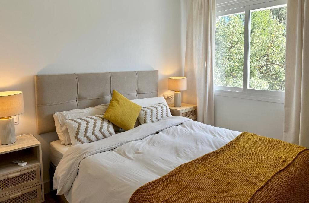 A neatly made bed with white bedding and a mustard-colored throw blanket, two bedside lamps, and a window with a view of trees.