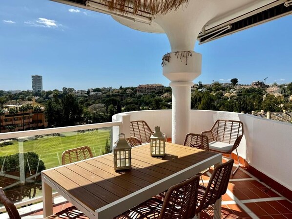 A balcony with a wooden table and wicker chairs, overlooking a cityscape.