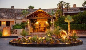 Un edificio con entrada de restaurante, rodeado por un jardín con diversas plantas y una llamativa luz circular decorativa.