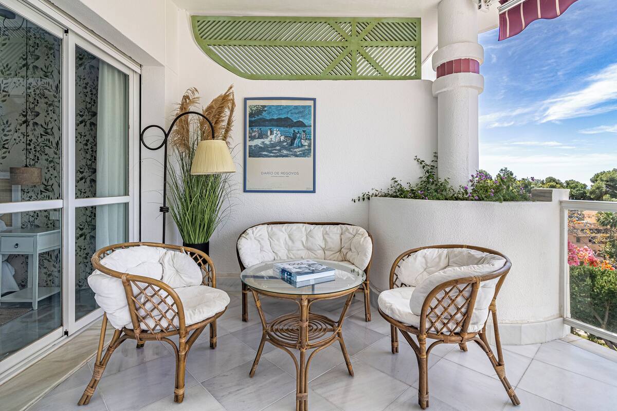 A balcony with wicker chairs, a glass table, a lamp, and a framed artwork.