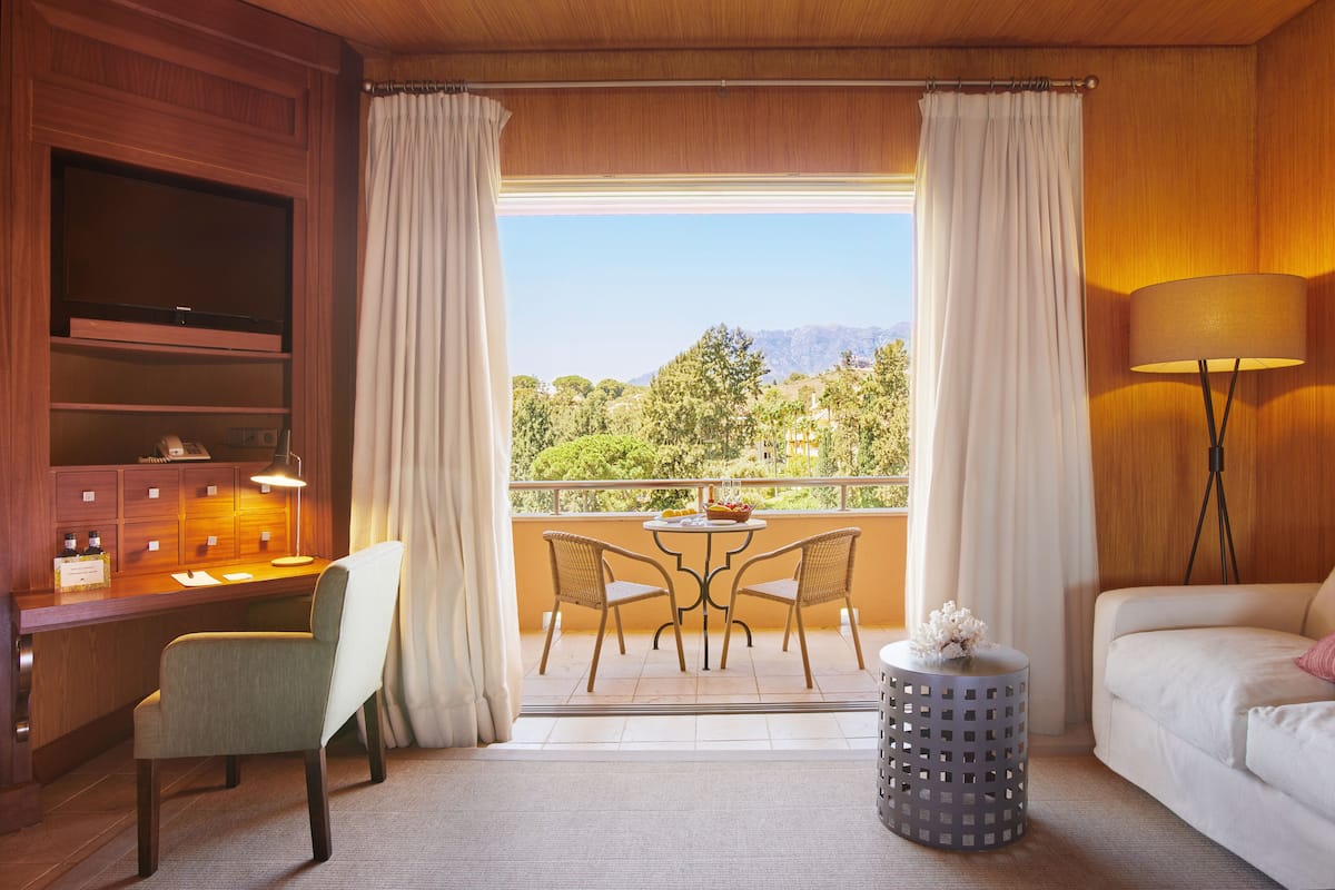 A hotel room with a desk, two chairs, a TV, a lamp, a round table with a flower, and a balcony with a view of trees and mountains.
