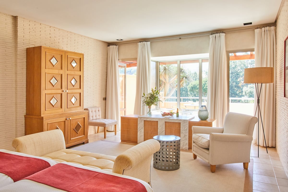 A hotel room with a large wooden wardrobe, a bed with red bedding, a desk with a chair, a small round table, and a lamp.