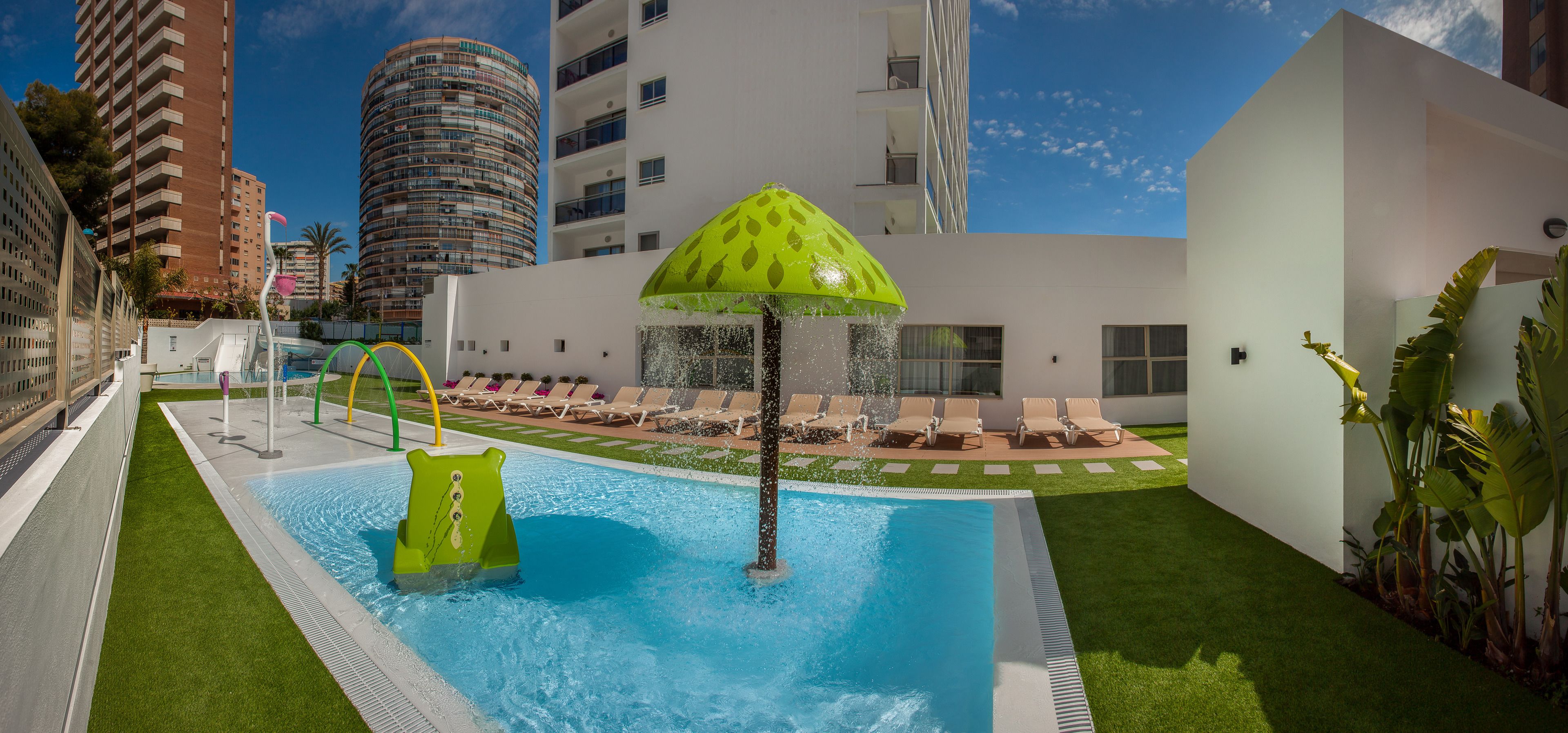 Una piscina con un tobogán de color verde, sillas de descanso y una sombrilla tropical.
