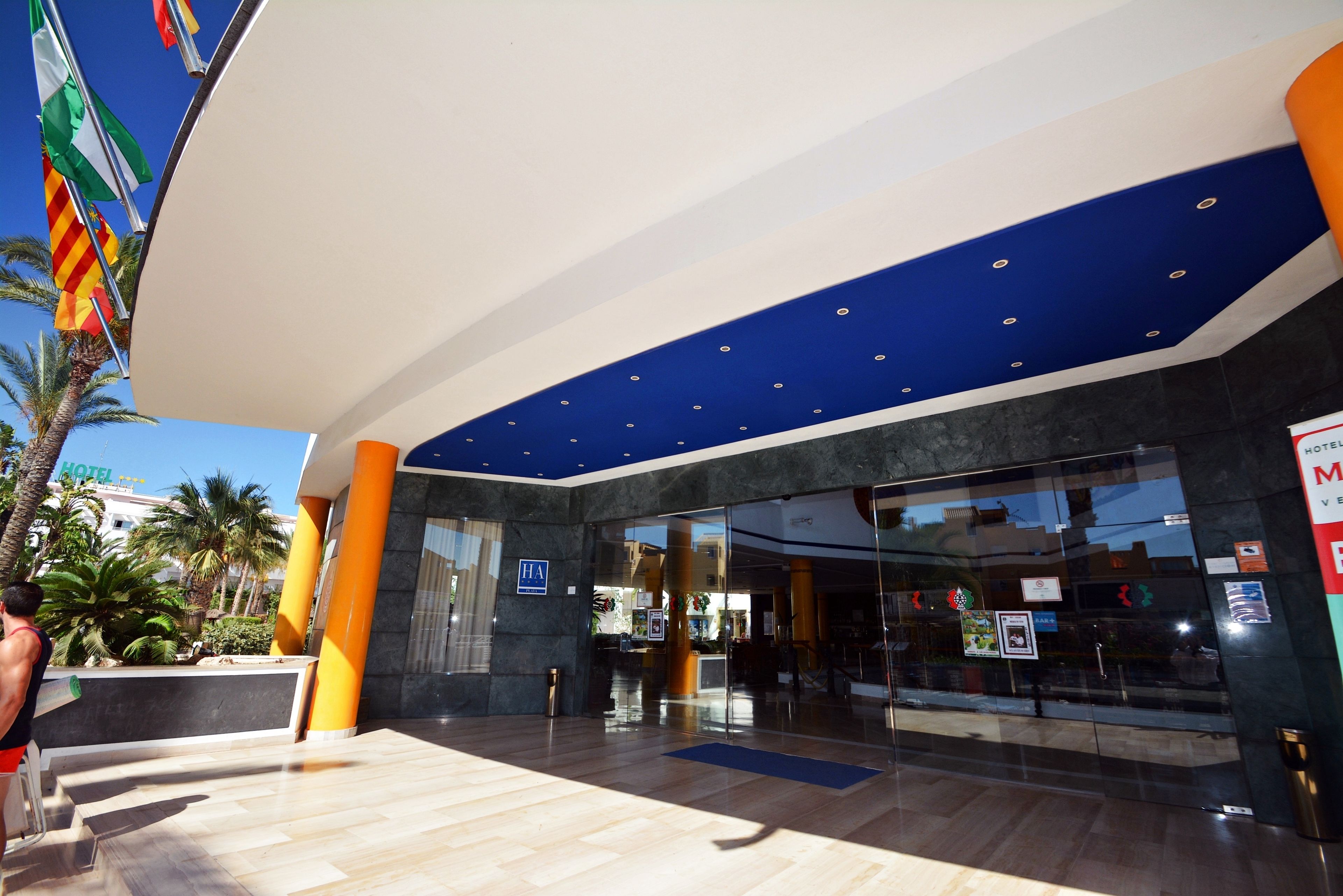 Fachada de hotel de diseño moderno, con techo azul, puertas de cristal y una palmera en el exterior.