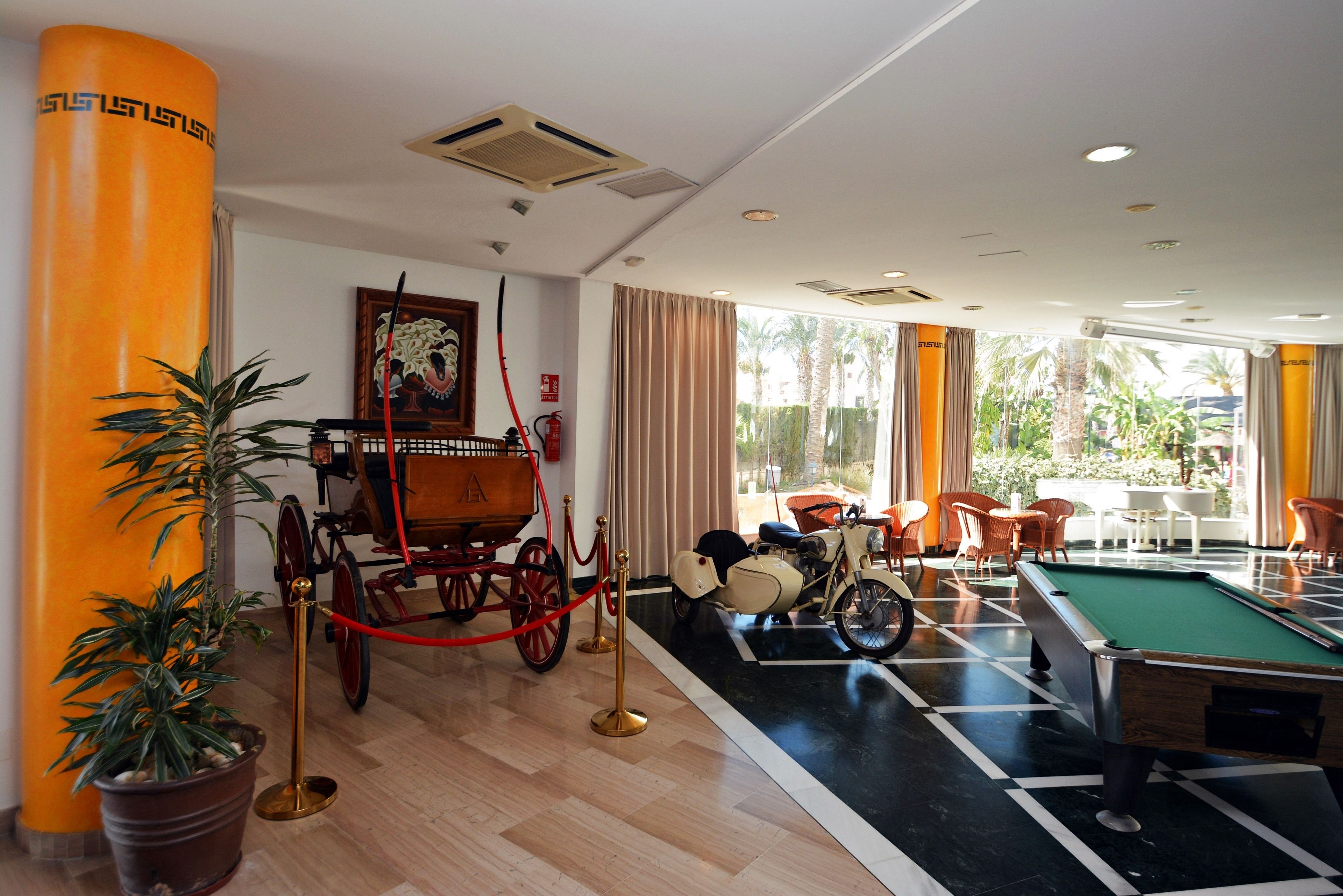 A billiards table, a vintage car, and a potted plant in a room with a wooden floor and large windows.