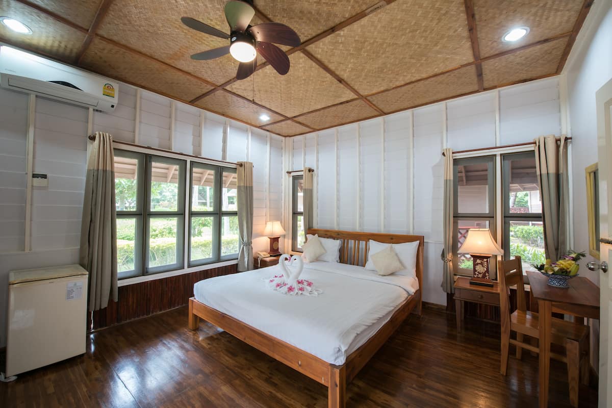 Un dormitorio con una cama de madera, un ventilador de techo, un escritorio con una lámpara y un refrigerador pequeño.