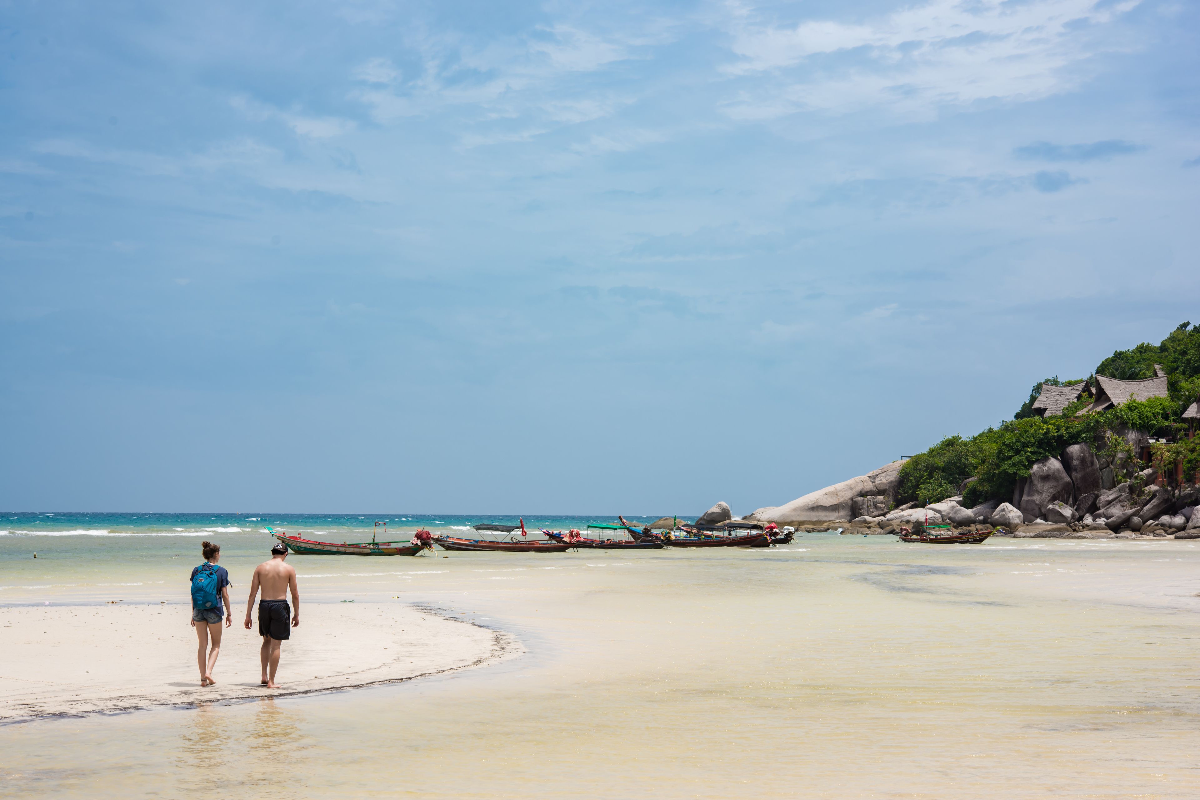 Una playa con una pareja caminando, barcos en el agua y acantilados rocosos.