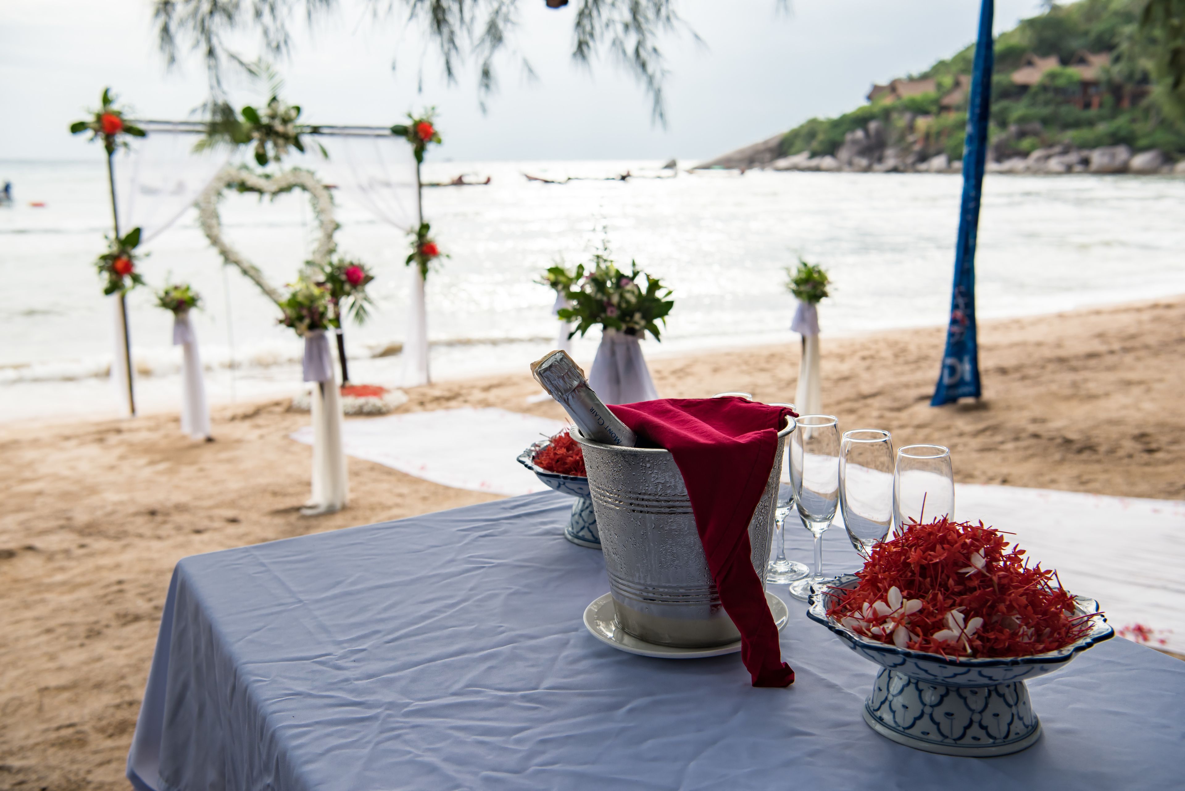 A table set with a blue tablecloth, a silver pitcher, a red napkin, and a bowl of red flowers.