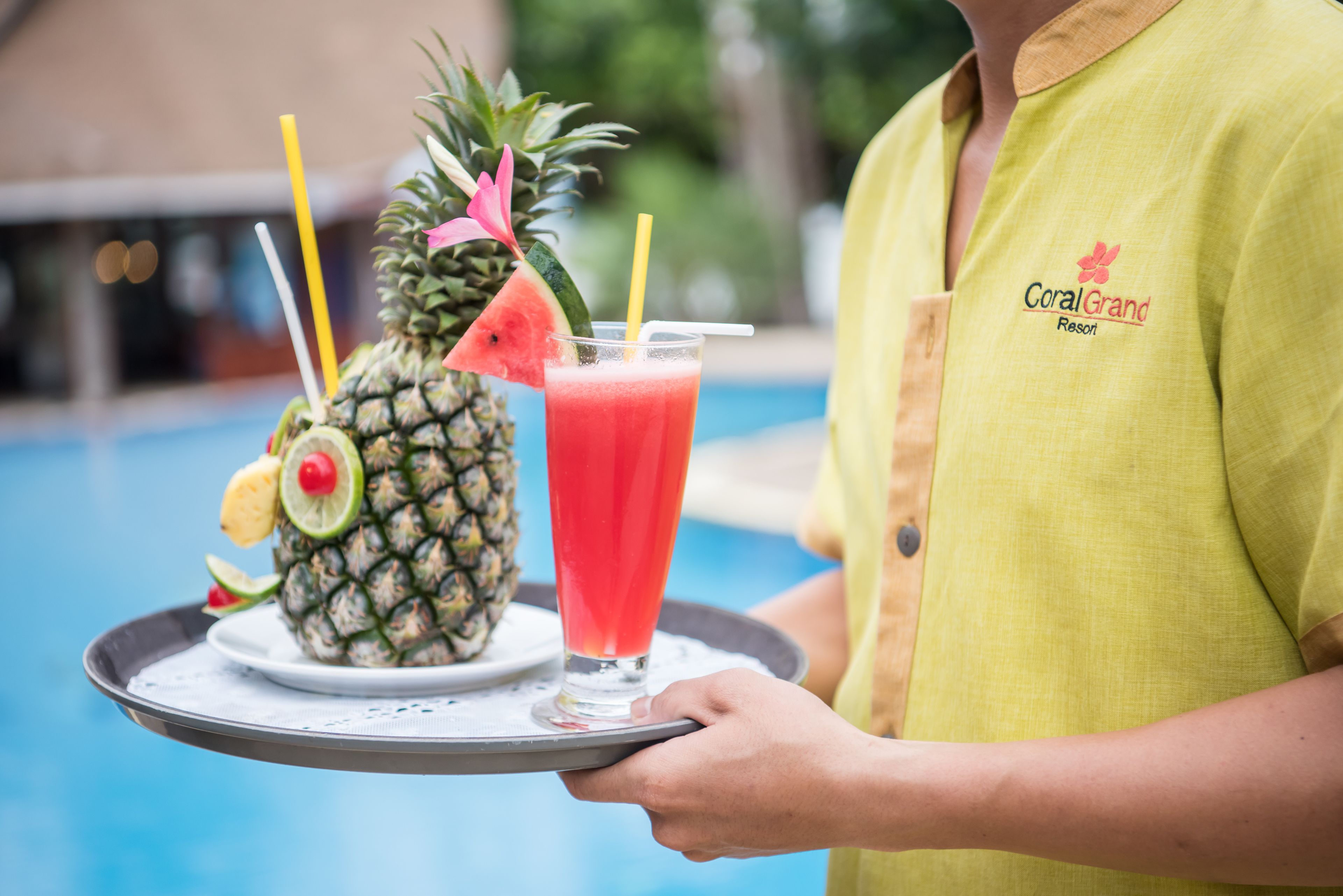 A person holding a tray with a pineapple and a glass of red drink, likely at a resort.