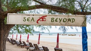 Una playa con sillas de descanso y sombrillas, un cartel que dice "SEA BEYOND" y barcos en el agua.