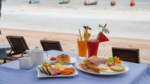 Una mesa de desayuno con diversas opciones de comida y bebidas, con vista a una playa y barcos al fondo.