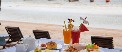 A breakfast table with a variety of food items and drinks, overlooking a beach with boats in the background.