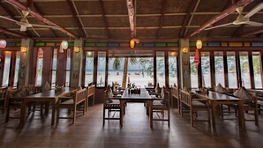 Un restaurante con mesas y sillas de madera, amplios ventanales con vistas a la playa y luces decorativas colgantes.