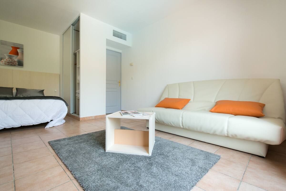 A modern bedroom with a bed, a white sofa with orange cushions, a small white side table, and a grey rug.