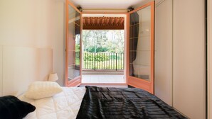 A bedroom with a bed, a nightstand, and a window with a view of a balcony and greenery.