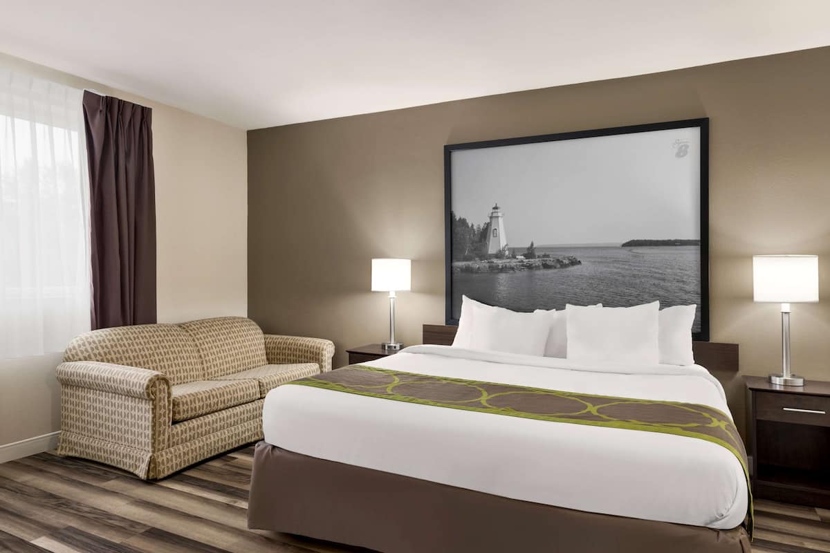A hotel room with a large bed, a sofa, two bedside tables with lamps, a large framed picture of a lighthouse, and a window with curtains.