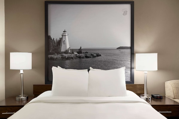 Habitación de hotel con una cama, dos lámparas de noche, un cuadro de un faro y una mesita de noche con un reloj.