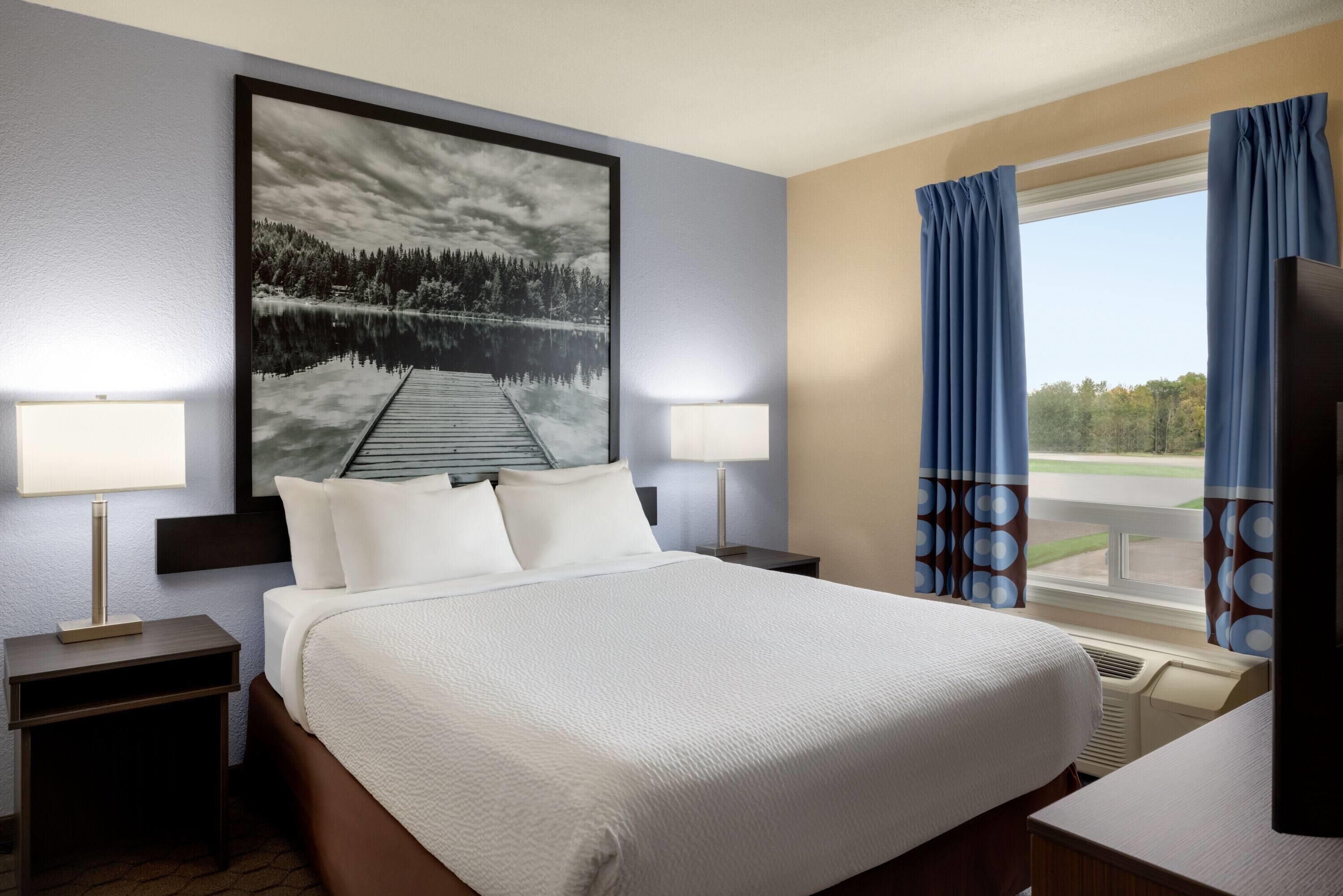 A hotel room with a large bed, two bedside lamps, a nightstand, a large window with curtains, and a painting of a lake.
