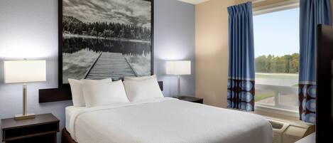 A hotel room with a large bed, two bedside lamps, a nightstand, a large window with curtains, and a painting of a lake.