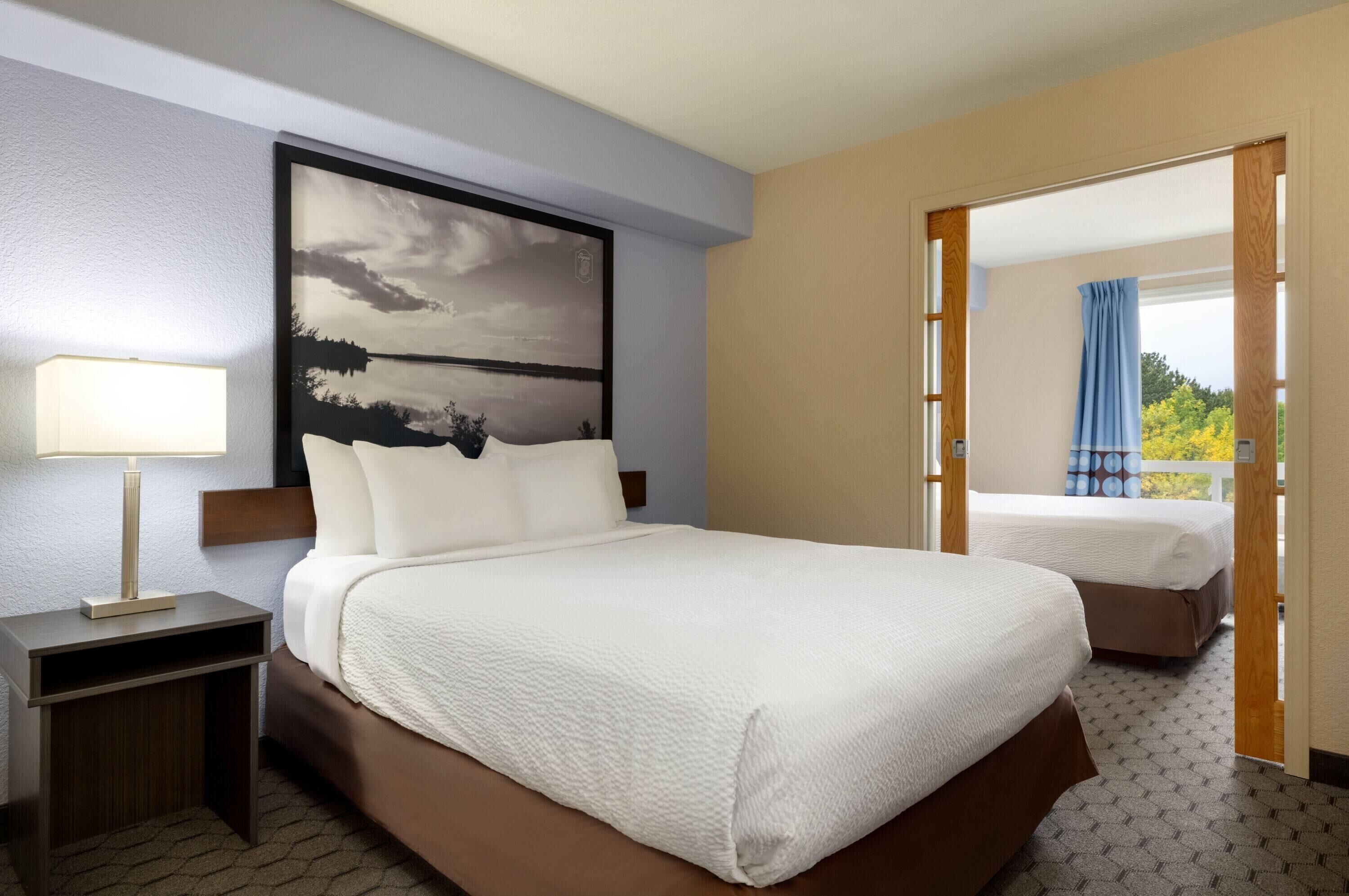 A hotel room with a large bed, a bedside table with a lamp, a view of the outdoors through an open door, and a painting on the wall.