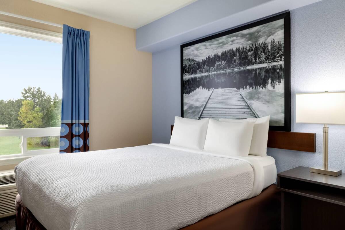 Une chambre d’hôtel avec un grand lit, une fenêtre avec des rideaux, un téléviseur fixé au mur et une table de chevet avec une lampe.