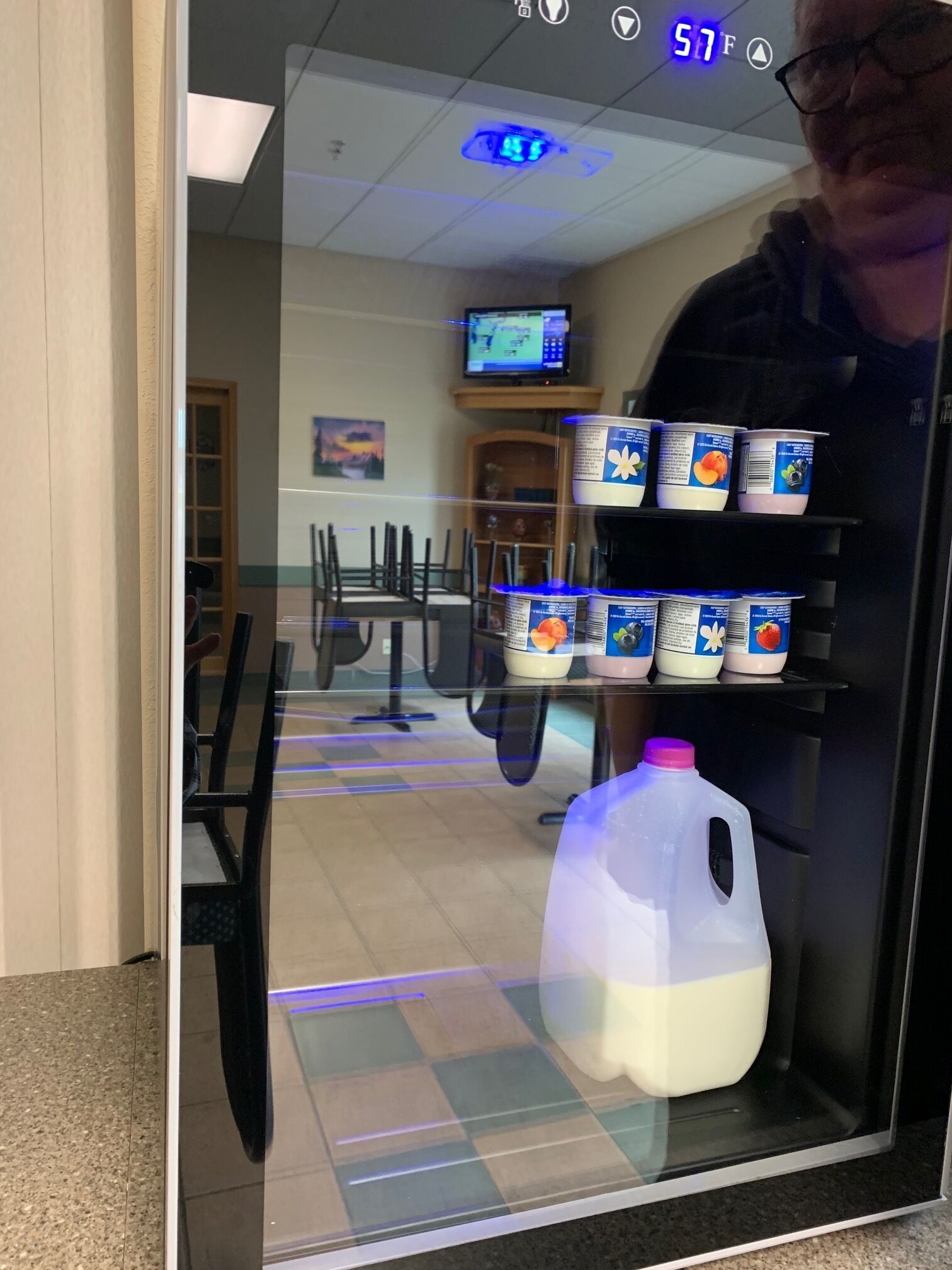 Un refrigerador lleno de diversos alimentos y una jarra de leche dentro de una habitación con piso de baldosas, un televisor y un cuadro en la pared.