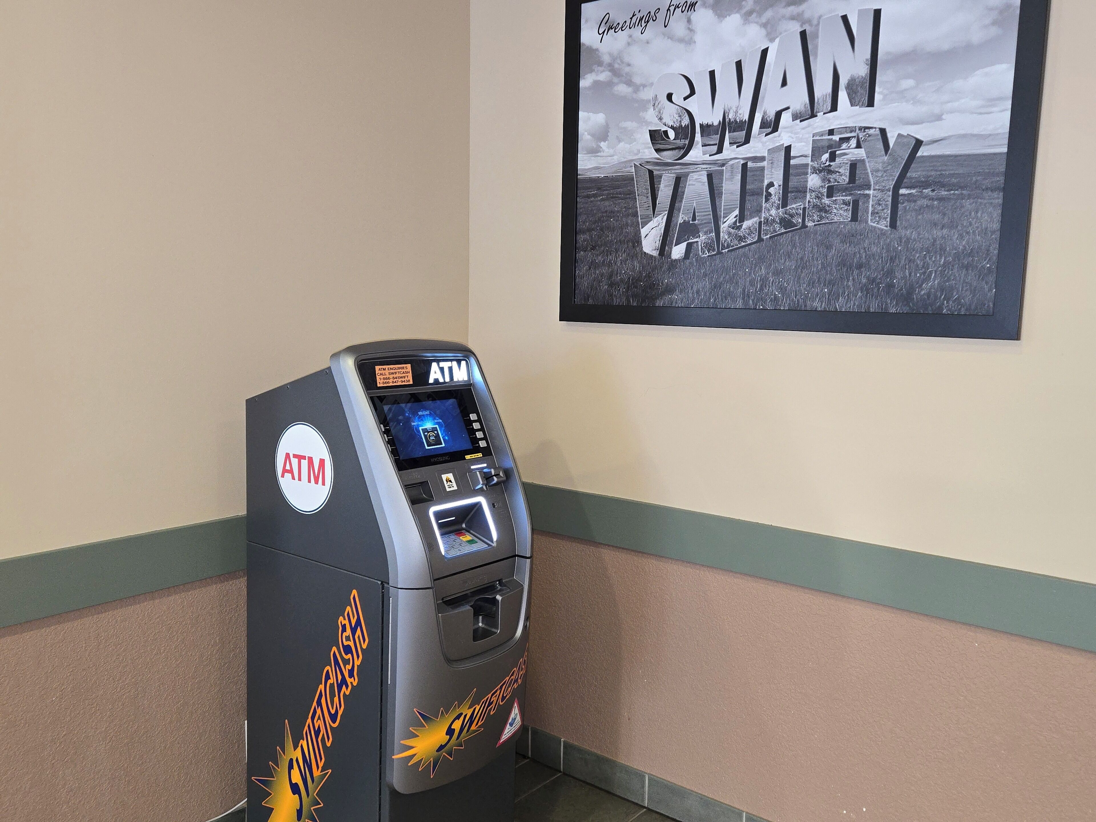 Ein Geldautomat mit 'SwifCash'-Logo, einem 'Swain Valley' Schild im Hintergrund und einer grün-beigen Wand.
