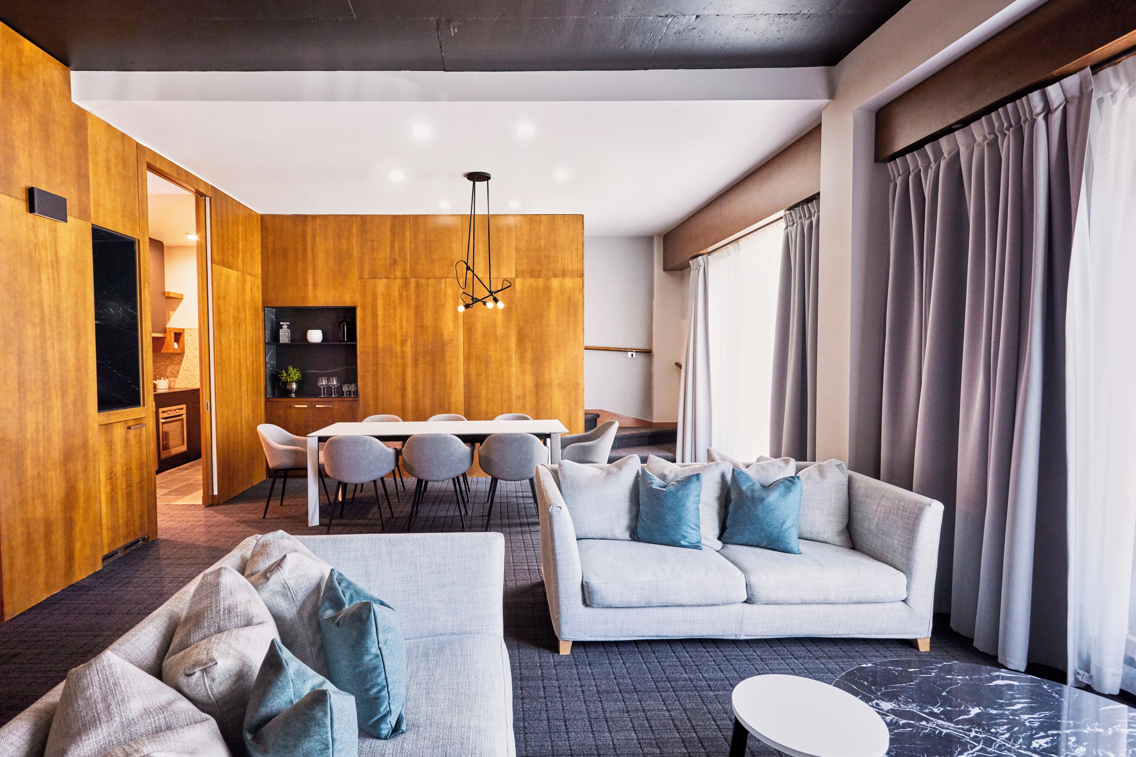Een moderne woonkamer met een bank, een eethoek met tafel en stoelen, een keuken op de achtergrond, en een groot raam met gordijnen.