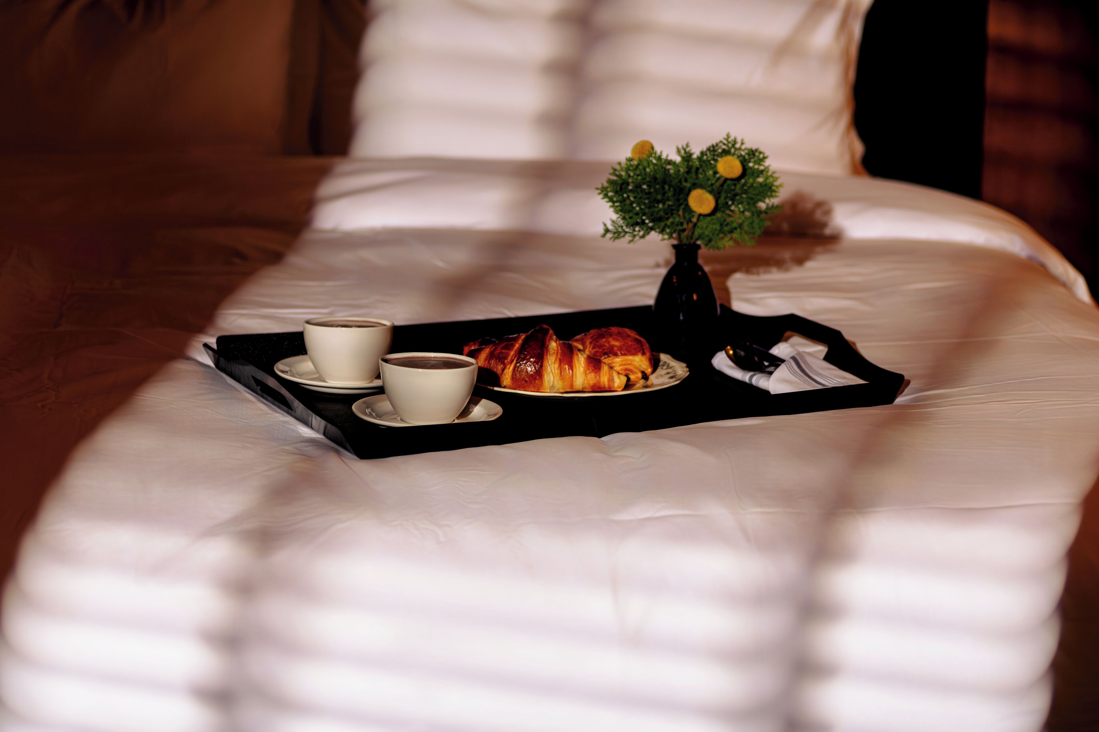Een net opgemaakt bed met een dienblad met croissants, een koffiekop en een kleine vaas met bloemen.