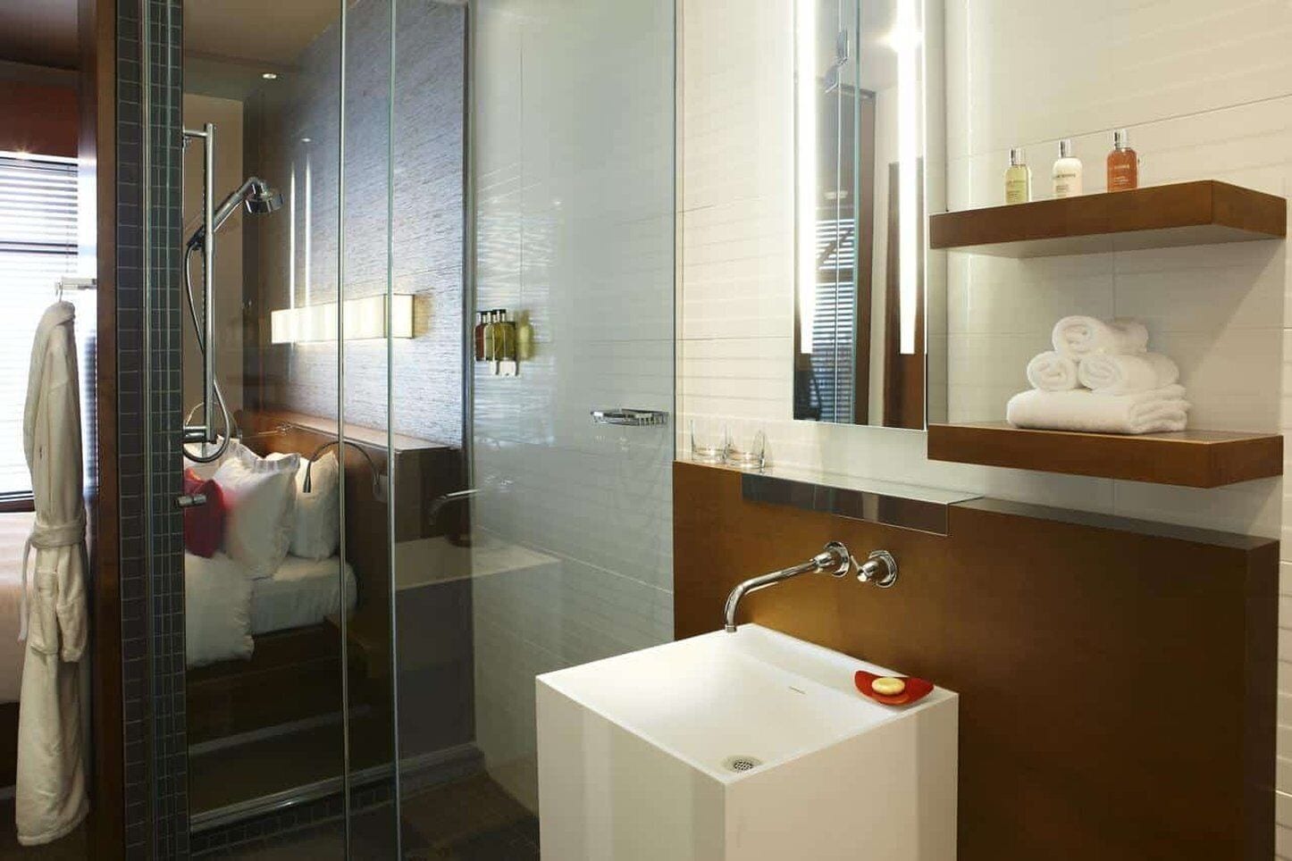 Een moderne badkamer met een douchecabine van glas, een wit wastafeltje en een spiegel erboven. Handdoeken en toiletartikelen liggen op een plank.