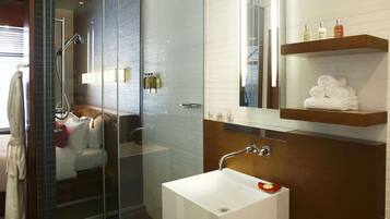 Een moderne badkamer met een douchecabine van glas, een wit wastafeltje en een spiegel erboven. Handdoeken en toiletartikelen liggen op een plank.