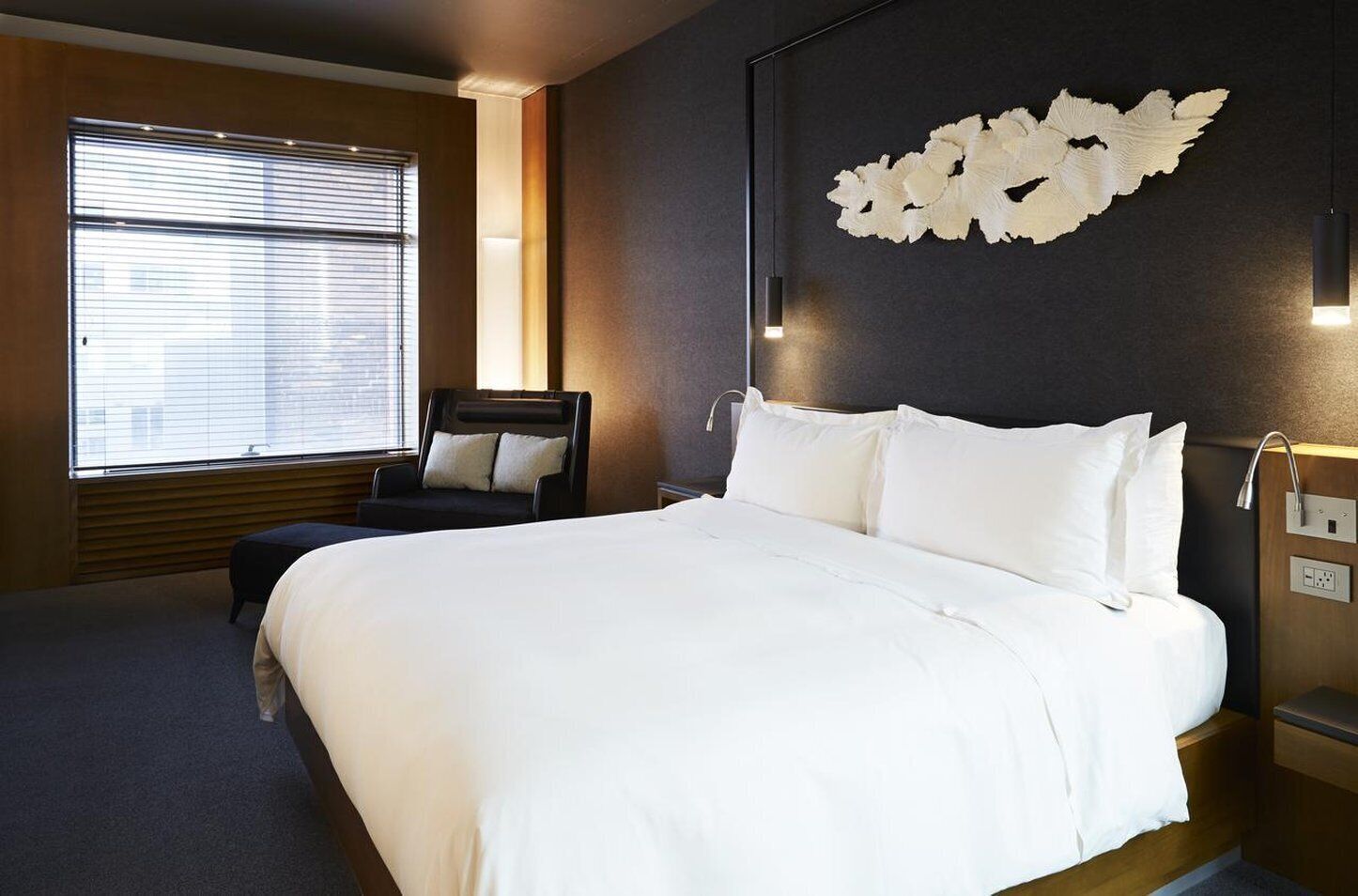 Ein modernes Hotelzimmer mit einem großen Bett, einem Sessel, einer Wandleuchte und einem Fenster mit Jalousien.